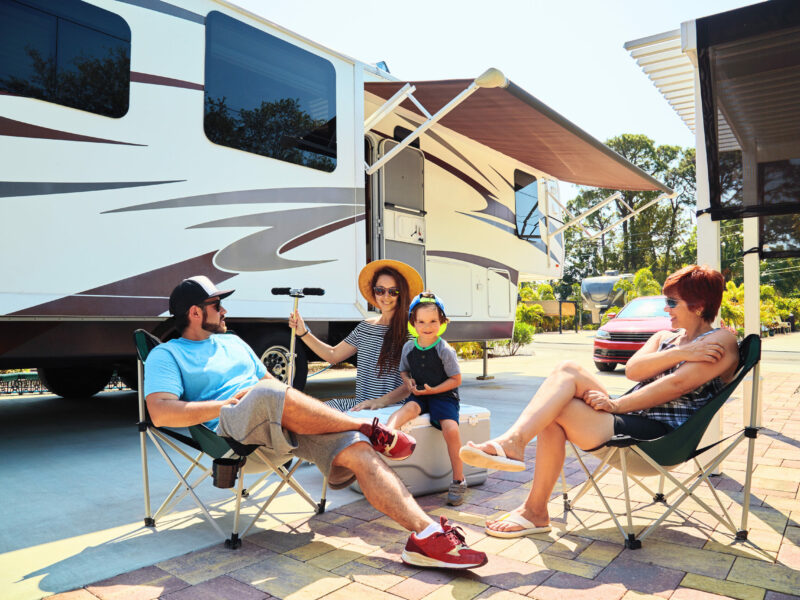 Mother,father,son and grandmother sitting near camping trailer,smiling.Woman,men,kid relaxing on chairs near car.Family spending time together on vacation near sea or ocean in modern rv park