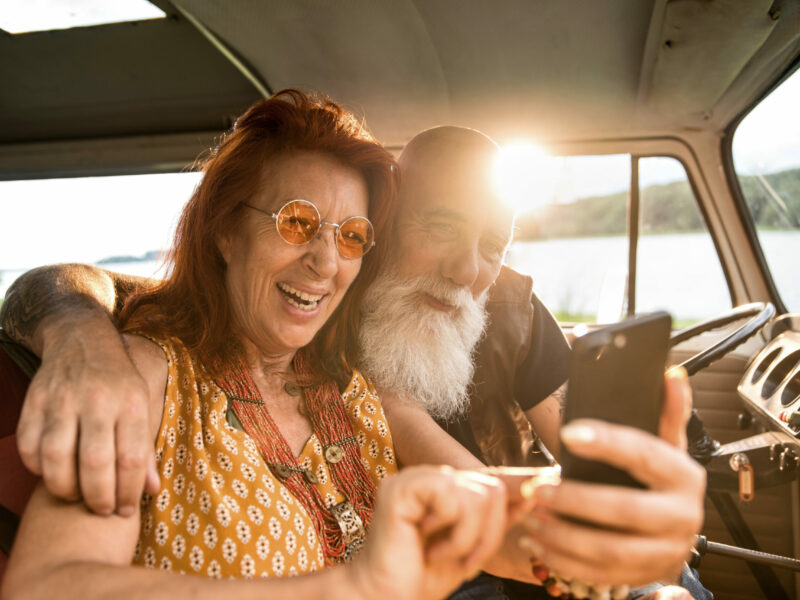 Portrait of an senior hipster couple using a smartphone inside their camper van parked near a lake. He's tattooed with a white beard, she's red hair.